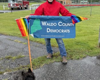 Andre Blanchard in a red raincoat holding the Waldo County Democrats banner and rainbow flags, with terrier Dugan