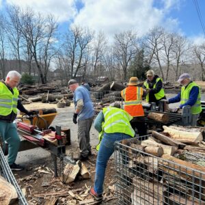 WCDC members in bright orange and green safety vests split and load firewood on an early spring day with bright blue sky and puffy white clouds
