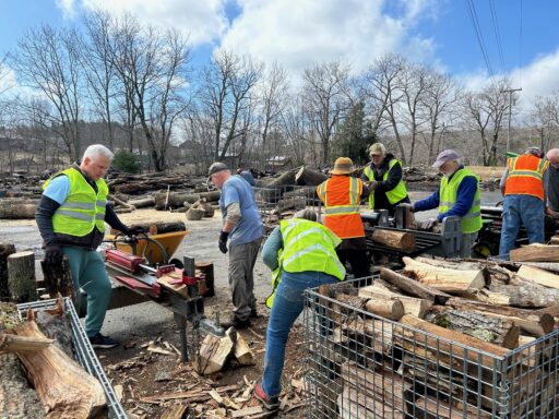 WCDC members in bright orange and green safety vests split and load firewood on an early spring day with bright blue sky and puffy white clouds