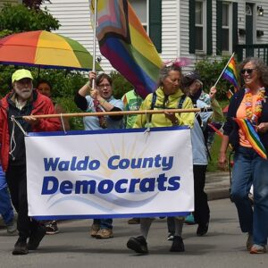 Volunteers marching in Belfast Pride Parade 2024, decked out in rainbox colors and carrying rainbow flags and the Waldo County Democrats banner.
