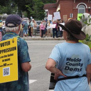 View of two protesters from behind, one with sign that reads "Voter Suppression is on the ballot in Maine, Vote NO Nov. 2025" and the other wearing a Waldo County Dems tee, facing a crowd of fellow protesters