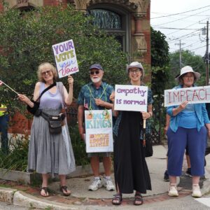 Group of protesters on Belfast street corner with signs reading "Your vote your voice," "empathy is normal," "No Kings," and "Impeach."