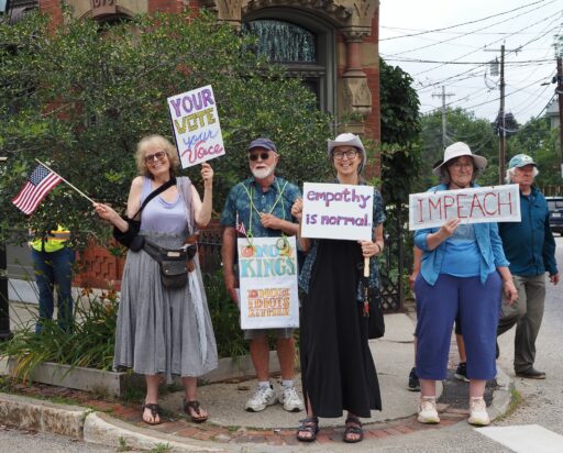 Group of protesters on Belfast street corner with signs reading "Your vote your voice," "empathy is normal," "No Kings," and "Impeach."