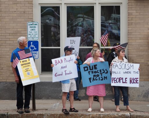 Protesters on the sidewalk in Belfast with signs reading "Voter Suppression is on the ballot in Maine," "BAD people need good trouble," Due process is not fascism," and "Fascism falls when the people rise."