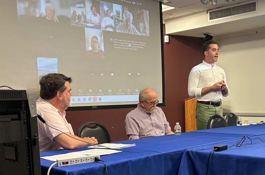 Senator Chip Curry and WCDC Chair Paige Zeigler listen as US Senate Candidate Jordan Wood addresses the Waldo County Democratic Committee on August 10, 2025. In the background is a screen showing Zoom participants.