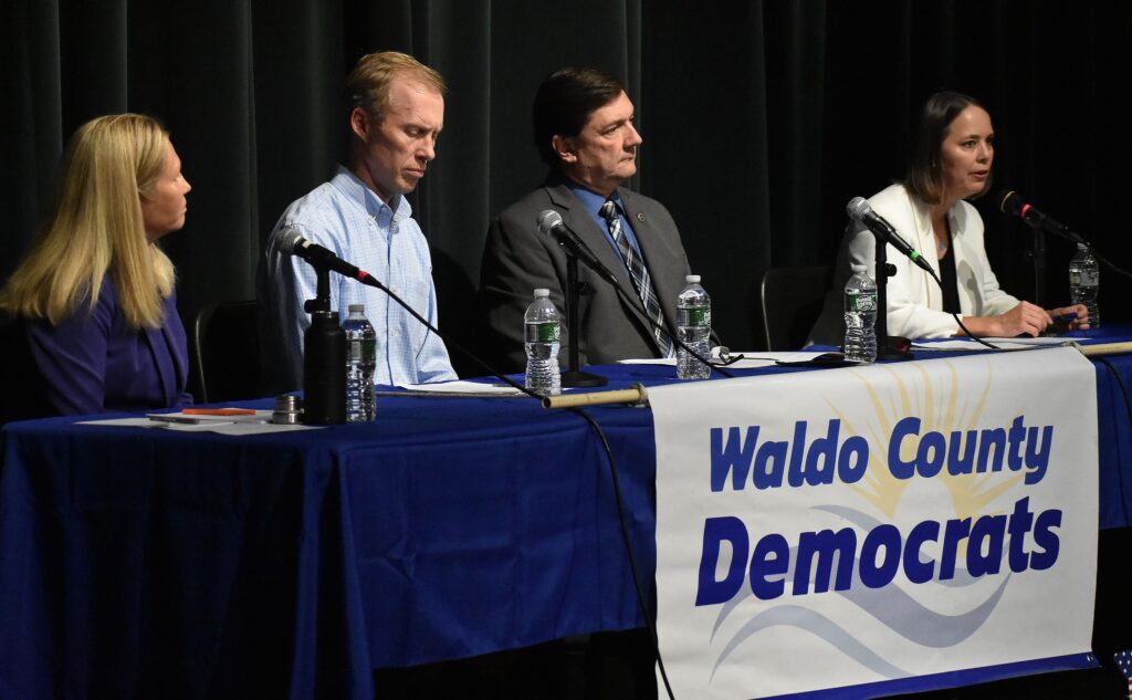 Four of the candidates (left to right, Hannah Pingree, Angus King III, Troy Jackson, and Shenna Bellows) are seated at a table with a blue and white Waldo County Democrats banner across the front.