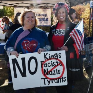 Rep. Jan Dodge, wearing a red tee and holding a US flag, is standing with a protester at the Oct. 18 No Kings Rally. The protester is wearing a blue tee that says RESIST and holding a sign that says No Kings Fascists Nazis Tyrants