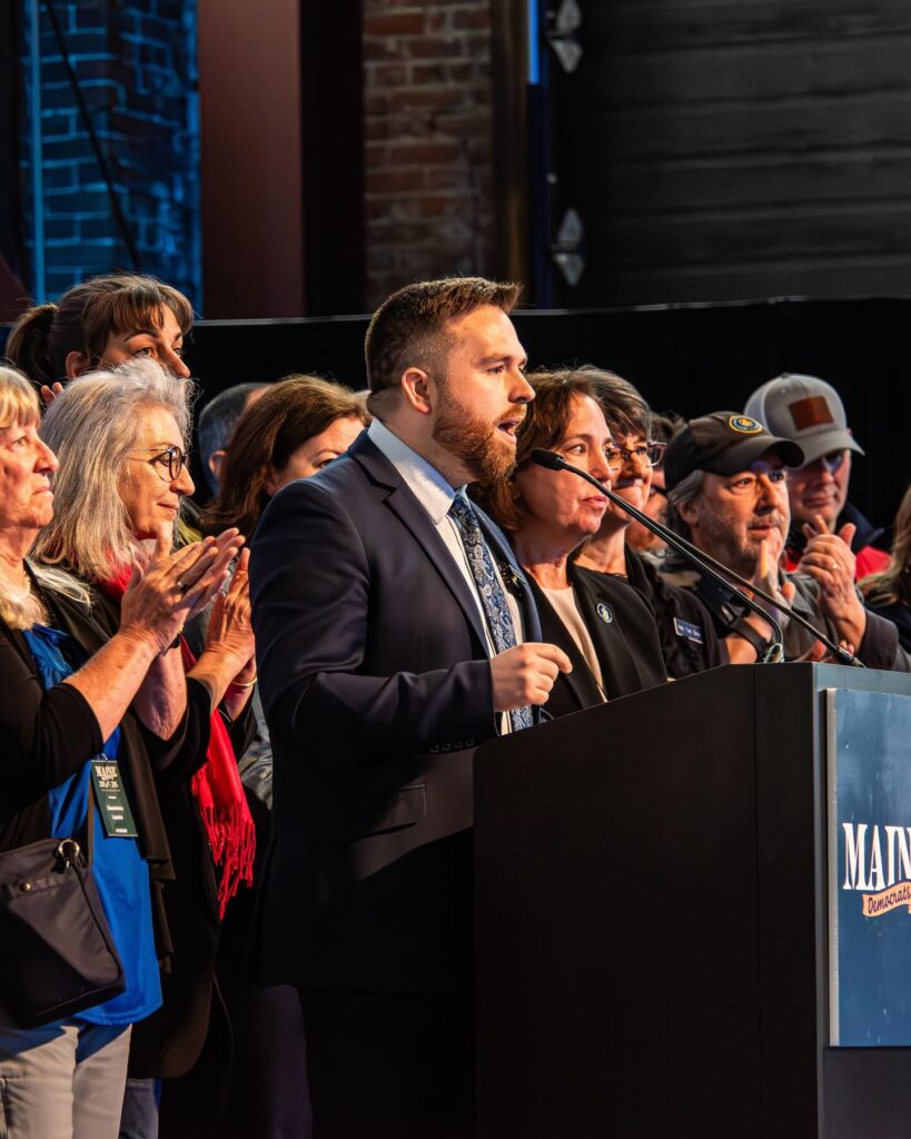 Ryan Fecteau speaks at the podium, surrounded by members of and candidates for the Maine House of Representatives.