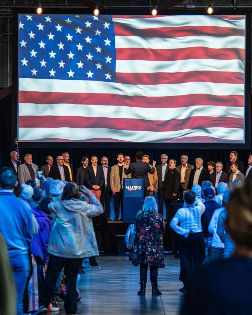 The Portland Gay Mens Chorus sings the national anthem below a waving flag shown on the big screen.