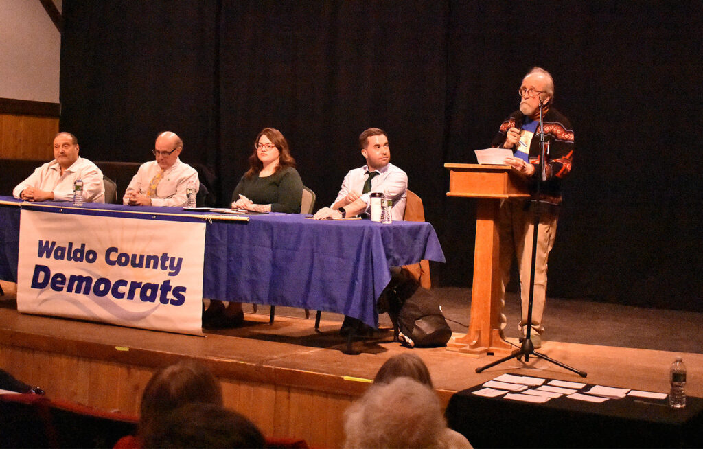 Candidates (l to r) Joe Baldacci, Matt Dunlap, Paige Loud, and Jordan Wood sit at a table with the Waldo County Dems banner across the front, with WCDC Chair Paige Zeigler standing at a lectern to their left. 