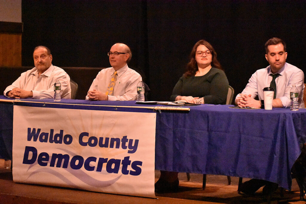 Candidates sit with hands folded behind a table draped in blue with the Waldo County Democrats banner across the front.