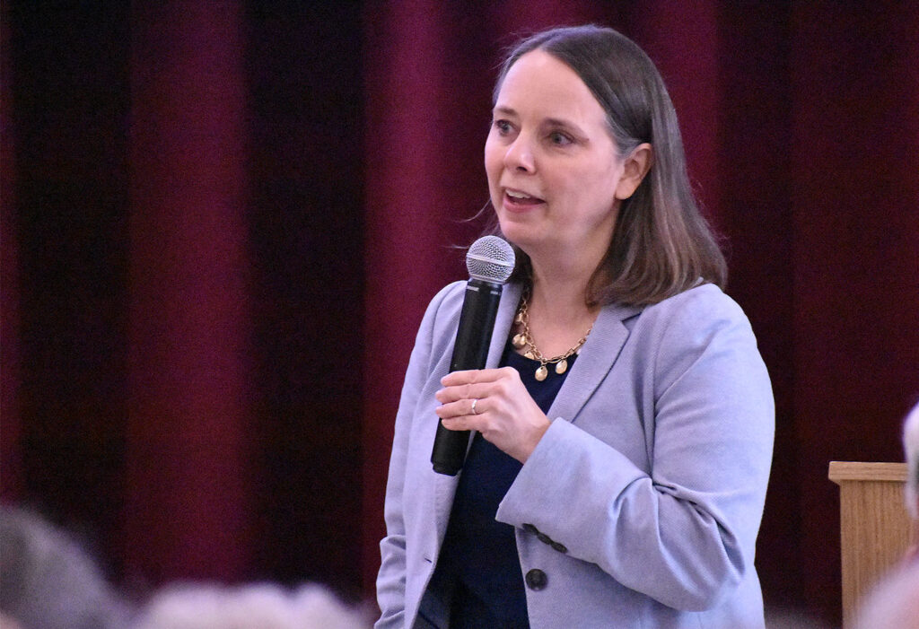 Secretary of State Shenna Bellows in a light blue jacket speaking into a microphone.