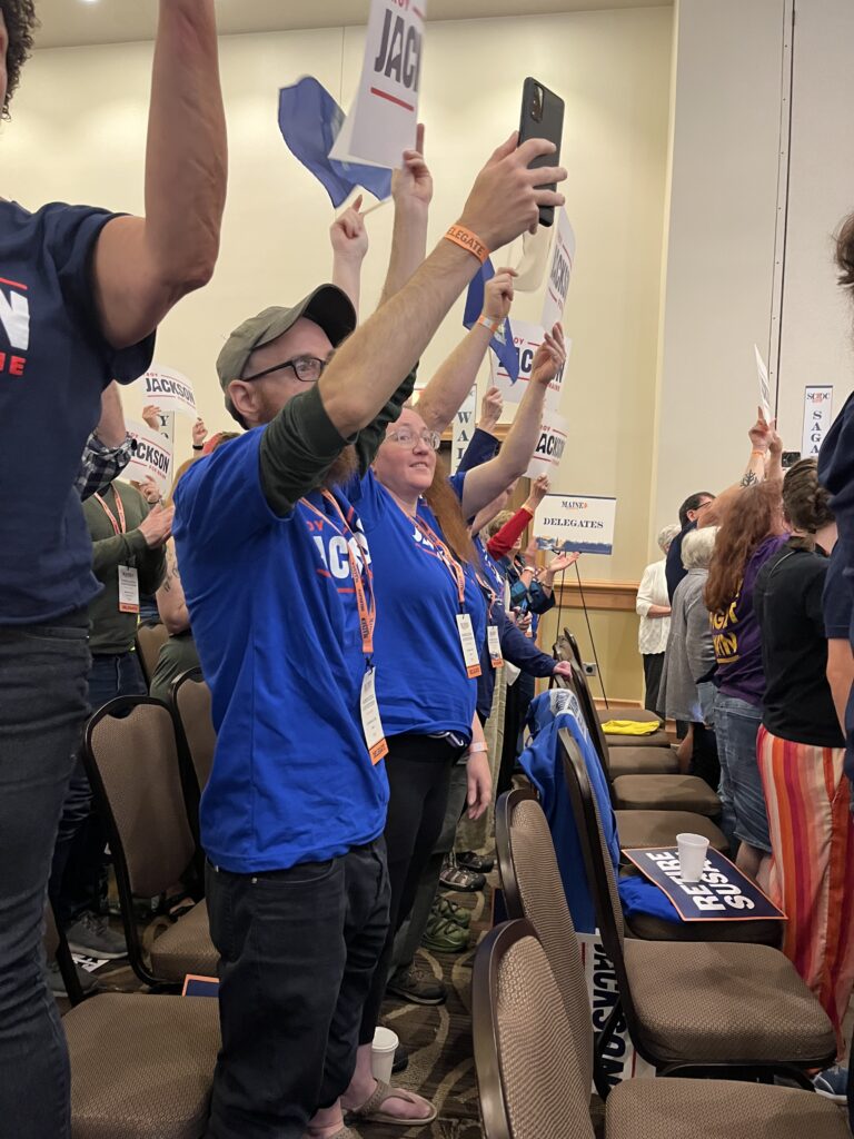 A row of cheering delegates in blue Troy Jackson shirts holding Jackson signs over their heads.