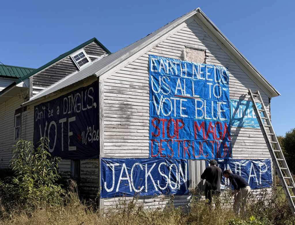 Big blue tarps and banners with white and red writing cover two sides of a clapboard building. They read - EARTH NEEDS US ALL TO VOTE BLUE, STOP MAGA DESTRUCTION, JACKSON DUNLAP, Don't be a DINGUS, VOTE 11/3/26

An extension ladder leans against the building and two people are on the ground attaching a banner.