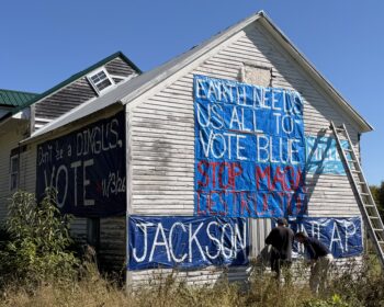 Big blue tarps and banners with white and red writing cover two sides of a clapboard building. They read - EARTH NEEDS US ALL TO VOTE BLUE, STOP MAGA DESTRUCTION, JACKSON DUNLAP, Don't be a DINGUS, VOTE 11/3/26